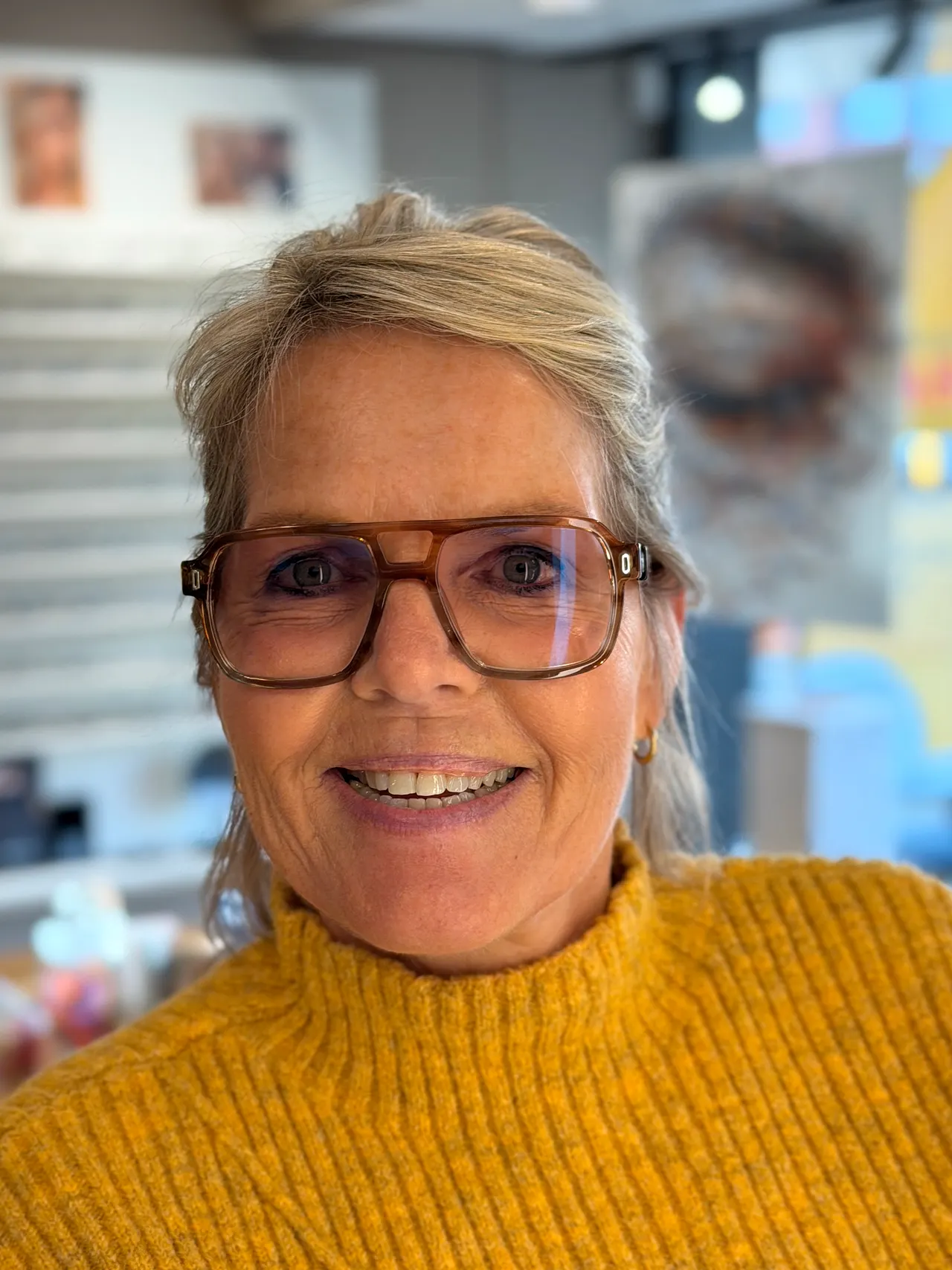 Portret van Gerda met bril en een okergele coltrui, lachend in de winkel.