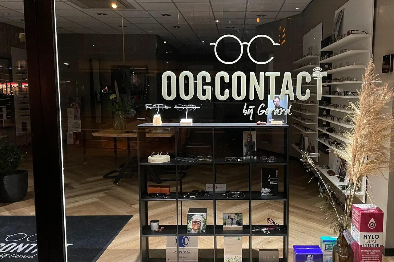 De winkel van buiten in de avond, met het verlichte logo Oogcontact by Gerard op de ruit.